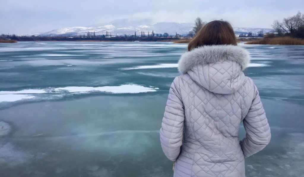 back of woman wearing light purple winter jacket near a frozen lake 