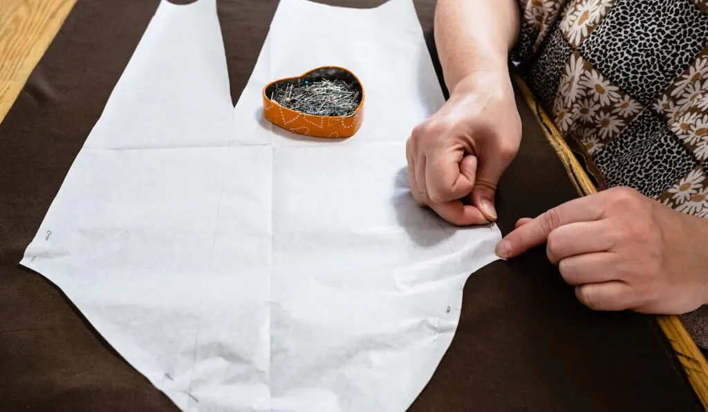 tailor fixes paper pattern layout of dress to brown fabric by pins on wooden table at home