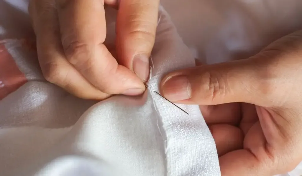 Hand of the seamstress using a needle and white thread to sew the seam on the shirt