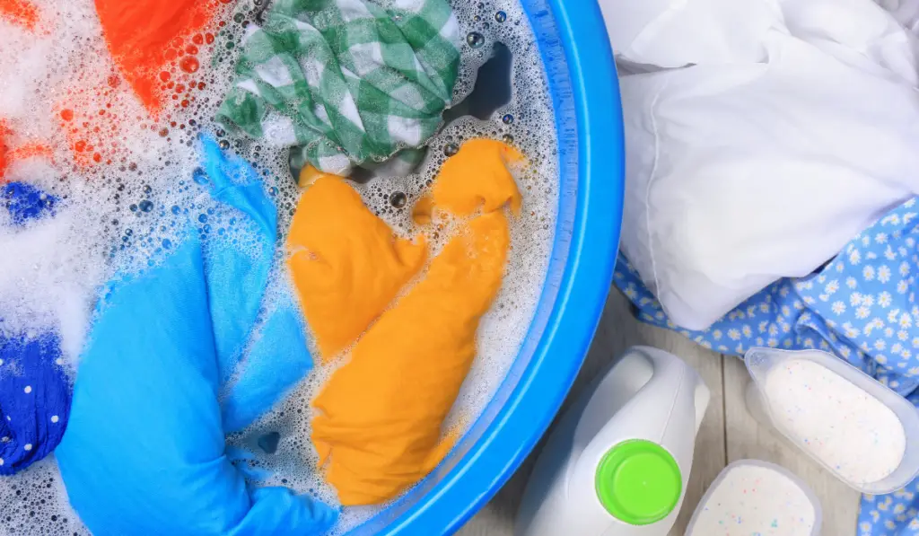 Basin with colorful clothes near bottle of detergent and powder on floor, flat lay Hand washing laundry
