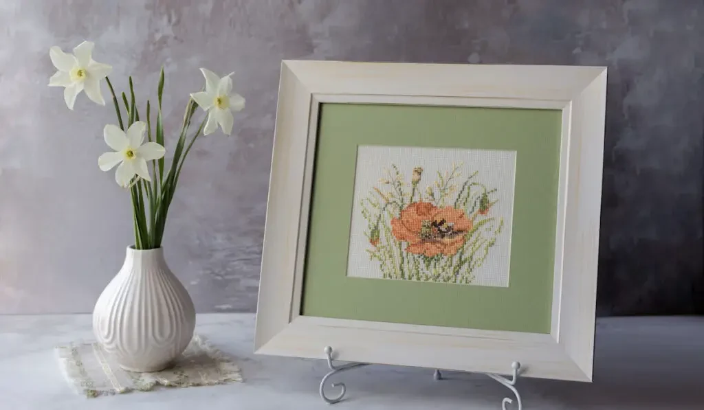 embroidered poppy flower in a wooden white frame next to a white flower vase