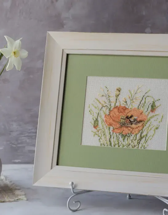 embroidered poppy flower in a wooden white frame next to a white flower vase